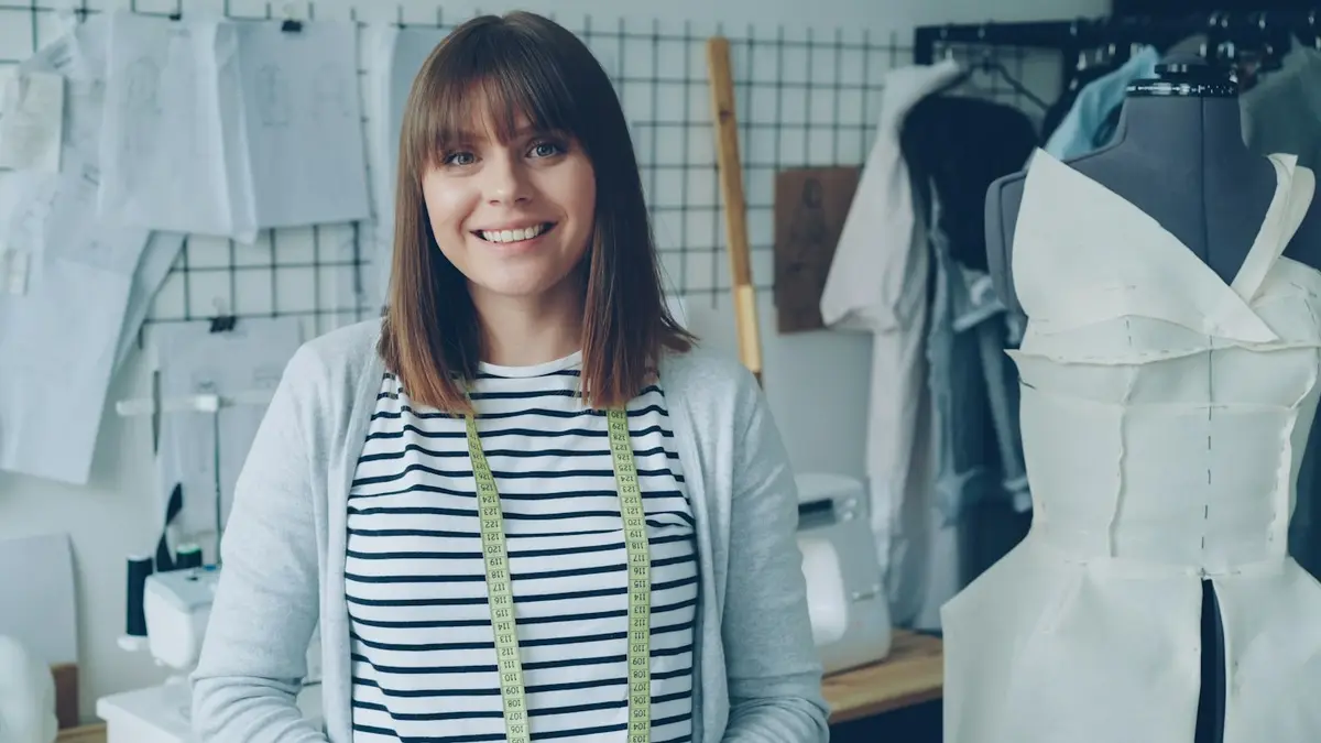 A dressmaker smiling in her workshop, a tape measure round her neck and a dress on a stand