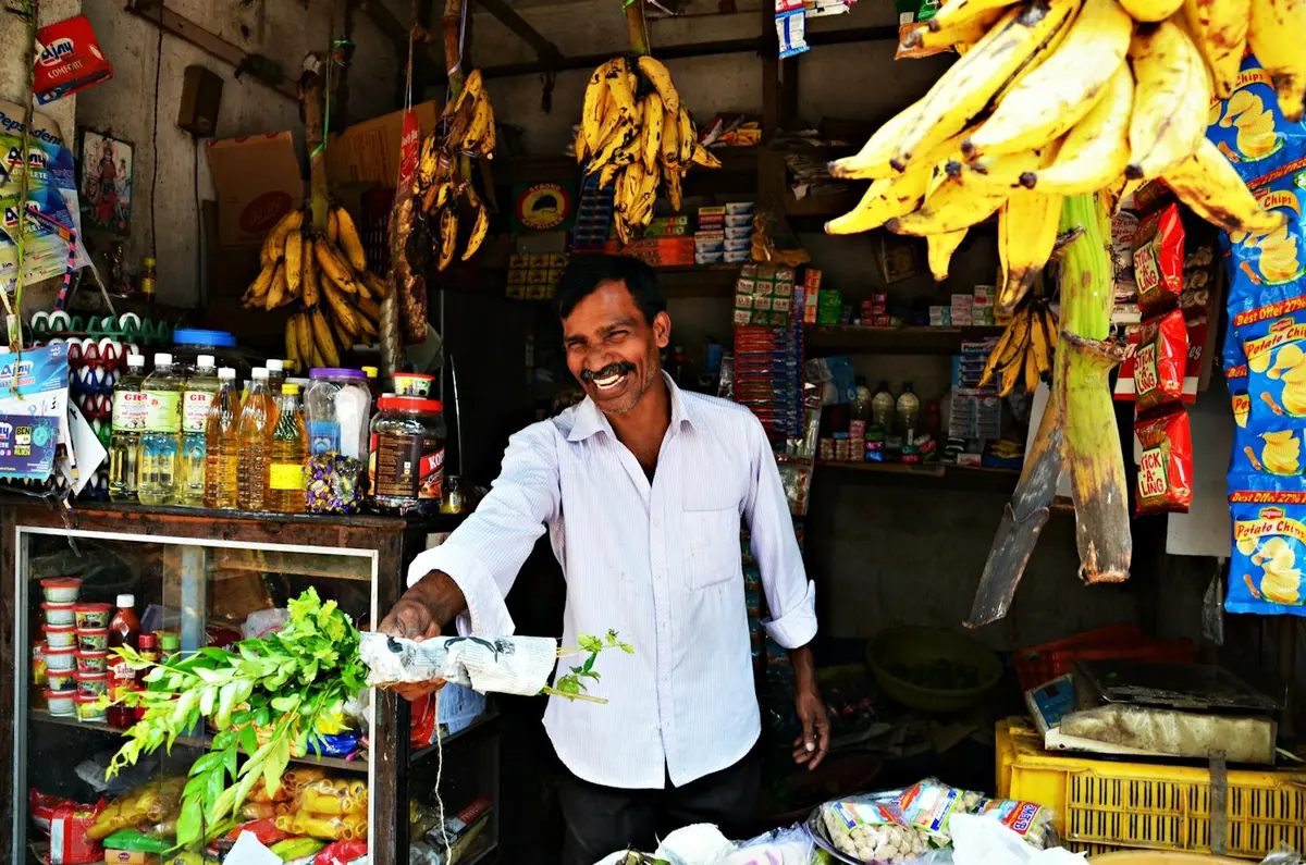 A shopkeeper smiling at the front of his grocery shop, holding out fresh curry leaves