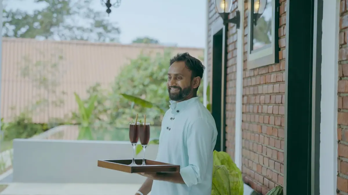 A smiling host carrying two drinks on a tray across a sunny terrace