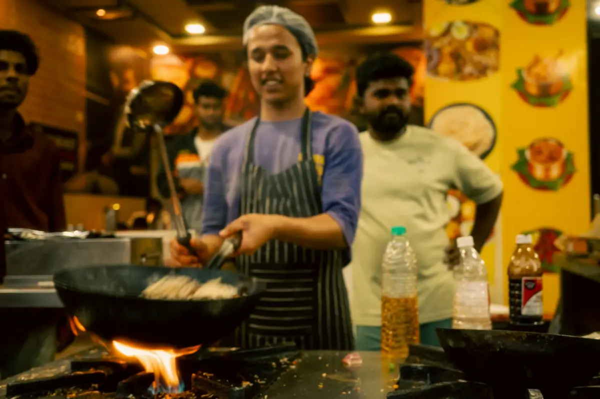 A cook tossing noodles in a wok over a flame in a busy restaurant kitchen